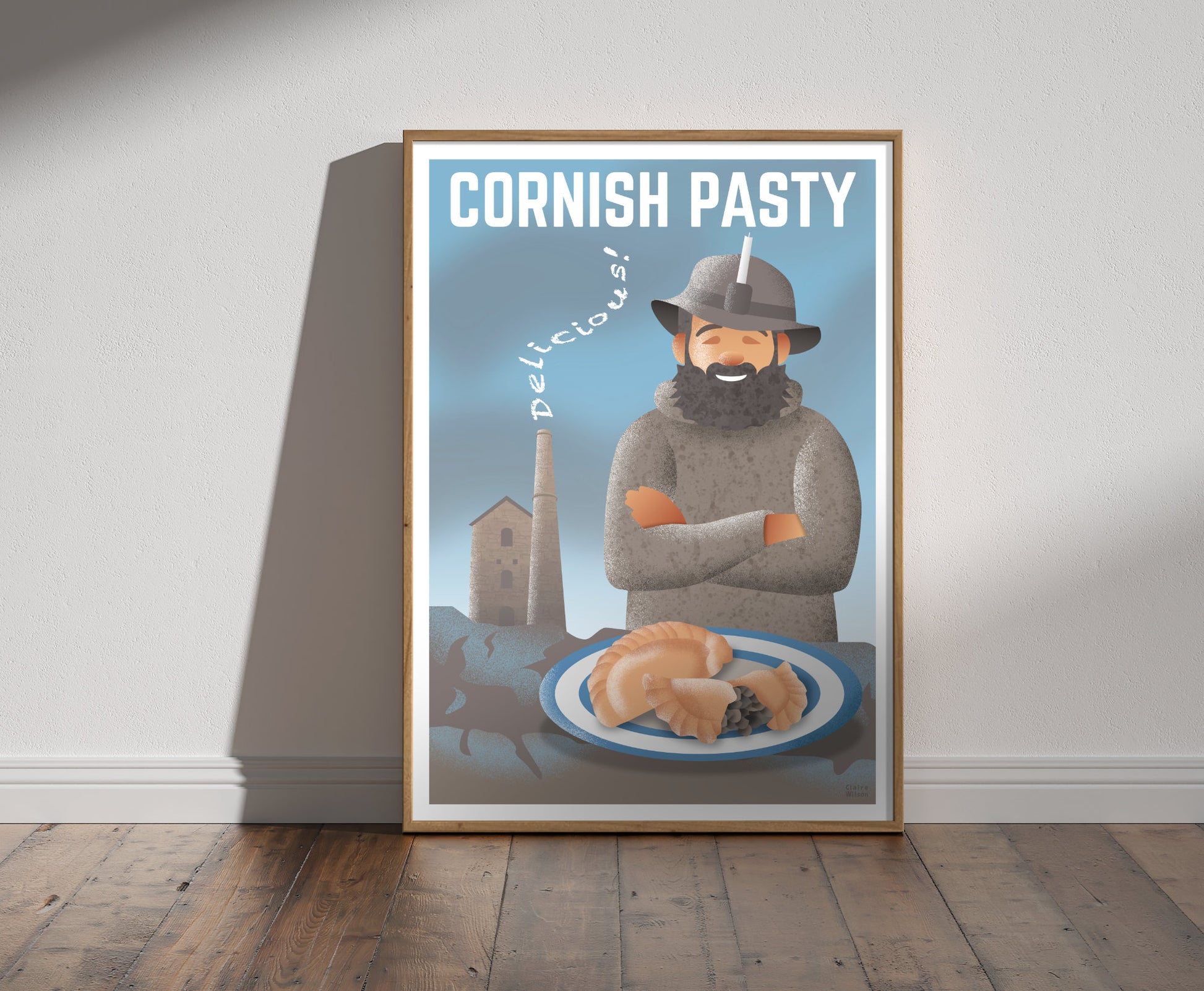 Framed illustration of a happy tin miner in light brown sweater, and a Cornish pasty, with a tin mine in background