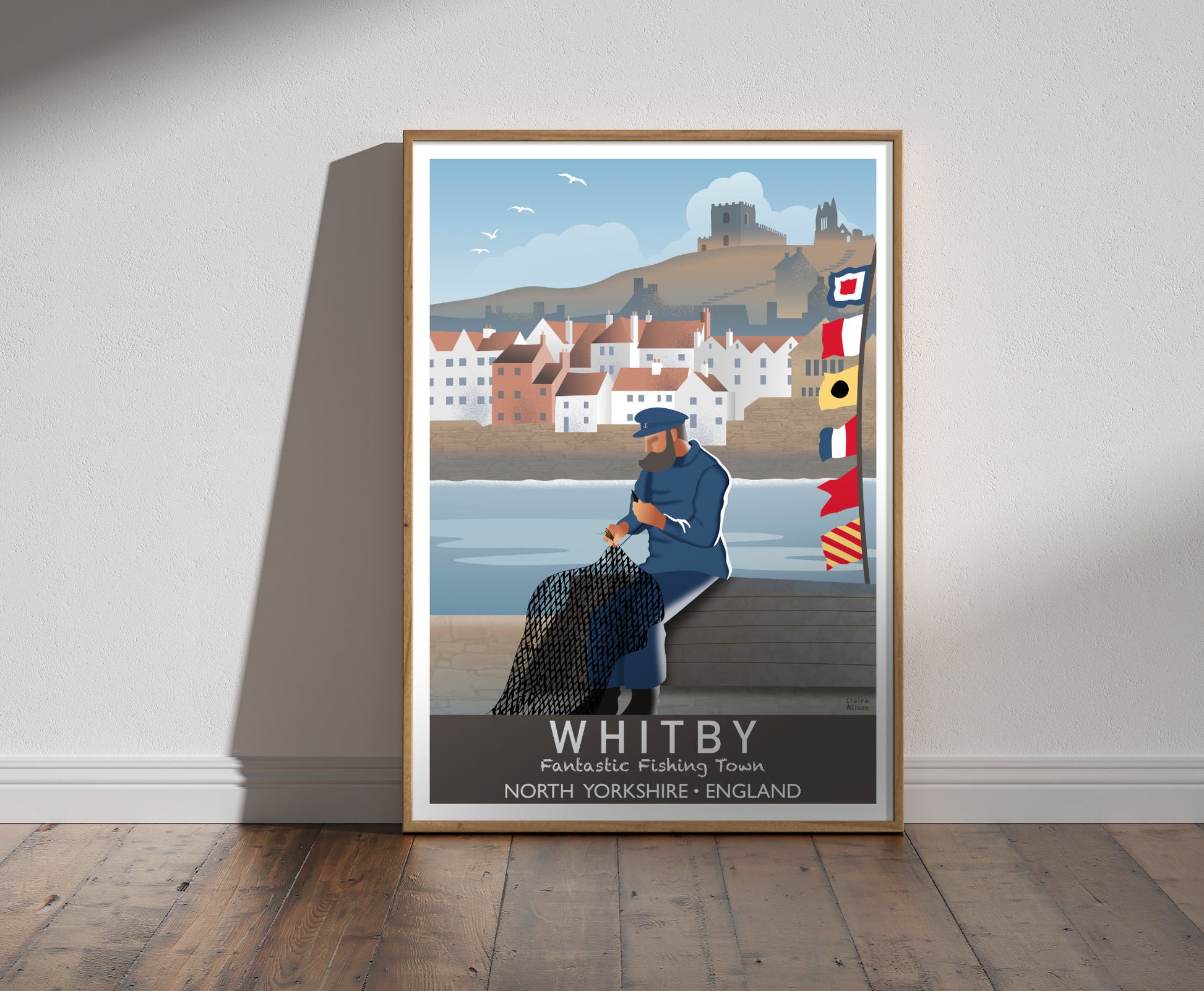 Framed illustration of a fisherman mending his nets by the harbour at Whitby, with nautical flags, old fishing cottages, a church and ruined abbey in the background 